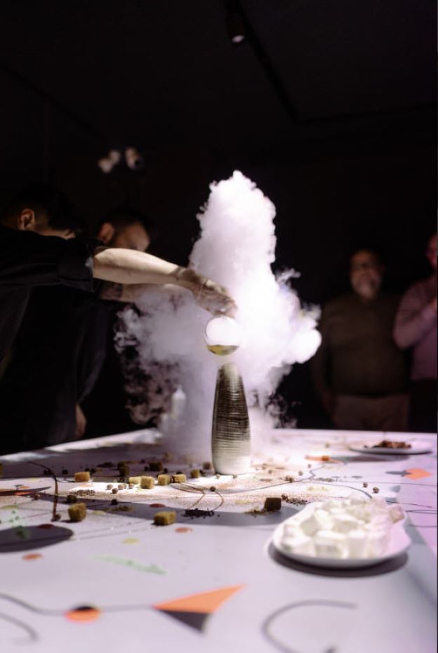 Una persona vierte líquido en un jarrón alto, haciendo que se eleve una nube de vapor, mientras se disponen postres y platos en una mesa decorada.